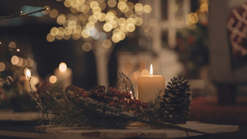A lit pillar candle beside a pine cone and a sprig of red berries on a wooden table with warm bokeh lights behind