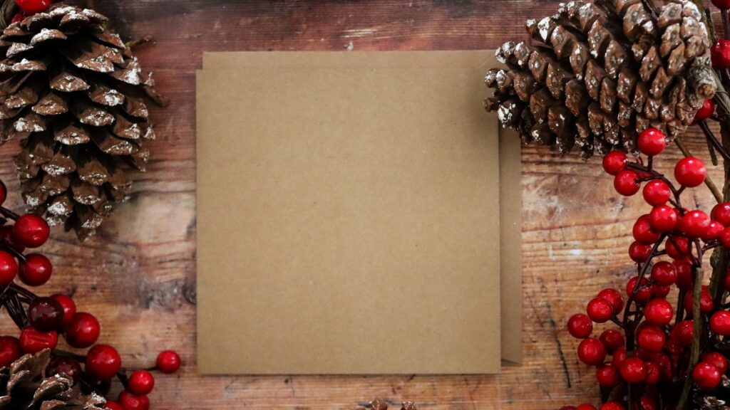A blank kraft-paper card and envelope on a rustic wooden table framed by pine cones and red berries