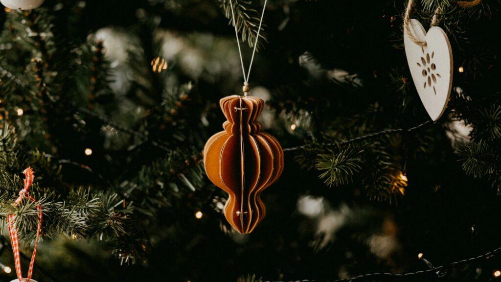 A copper paper ornament, a wooden heart and white bell decorations hanging on a dark green Christmas tree with warm lights