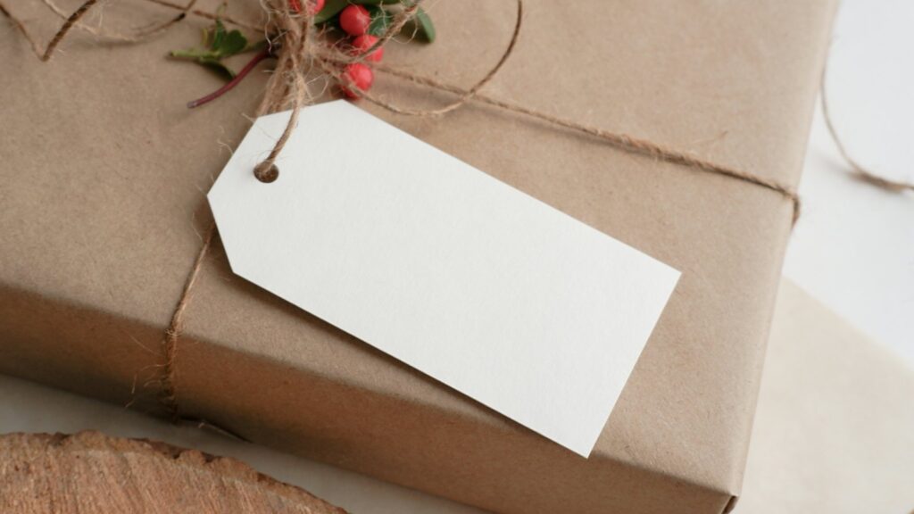 A gift wrapped in brown kraft paper and tied with jute twine, with a sprig of red berries and a blank white gift tag