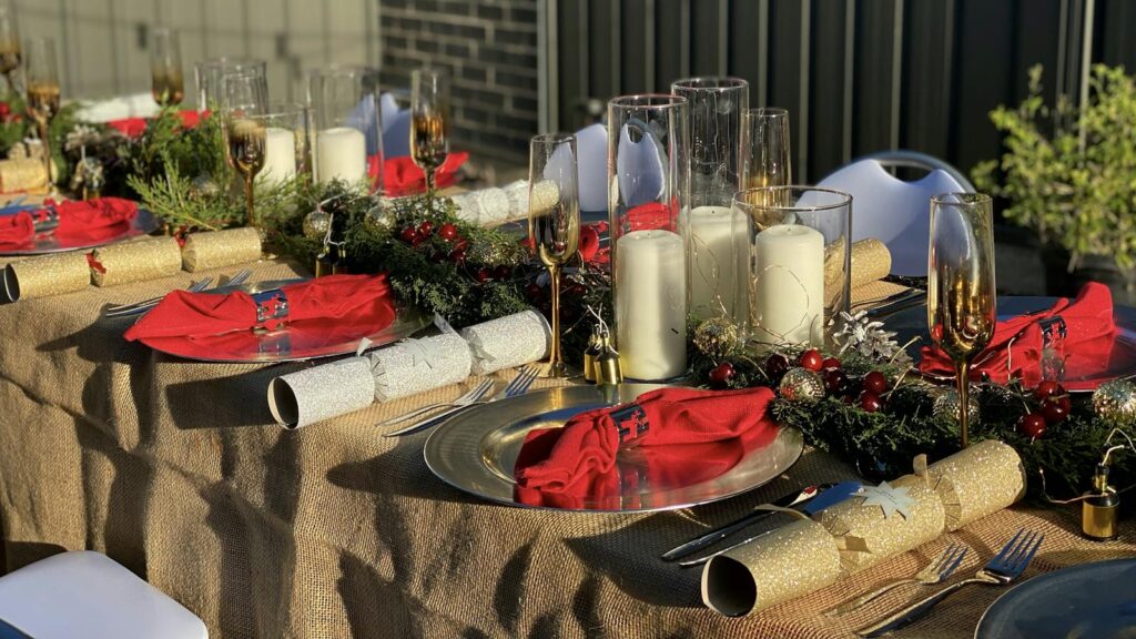 A long festive table set with red napkins, gold crackers, candles in glass and pine garlands on a burlap cloth