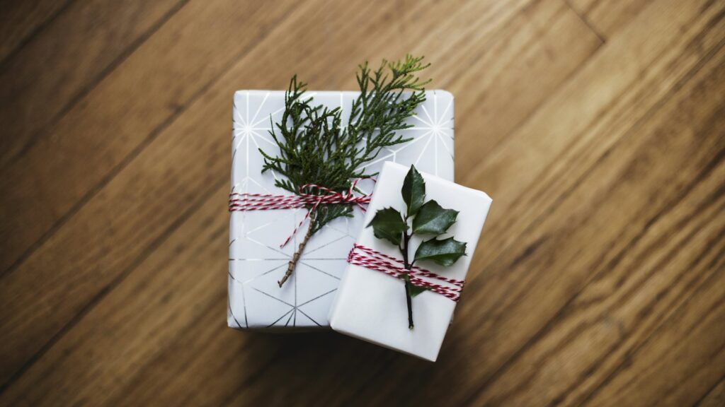 Two small gifts in white paper tied with red and white twine and topped with sprigs of greenery on a wooden floor