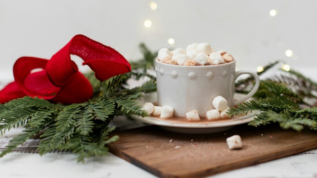 A white cup of hot chocolate with marshmallows on a wooden board beside pine branches and a red bow