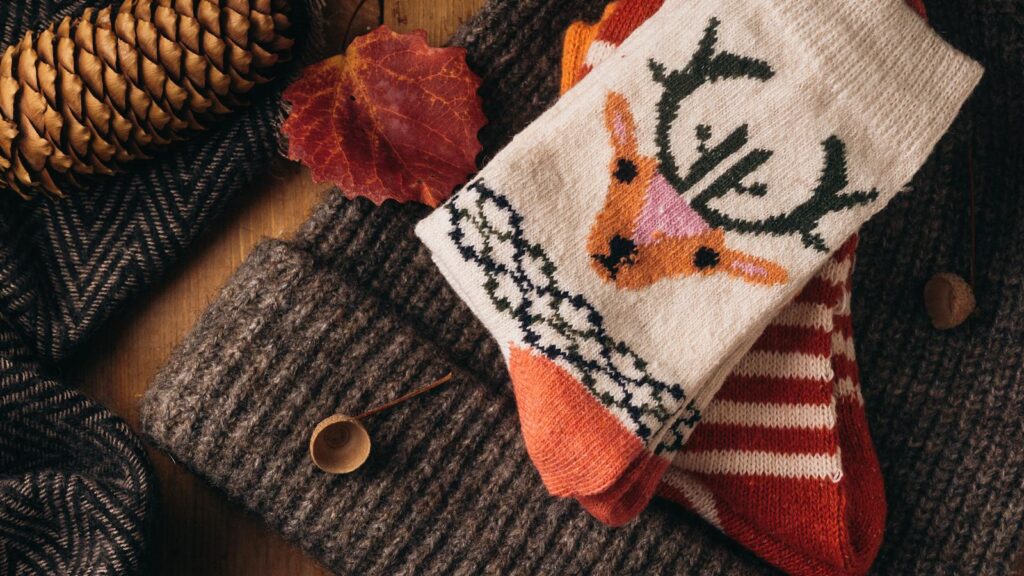 Patterned wool socks on a knitted hat beside a lit candle, pine cones and autumn leaves on a wooden table