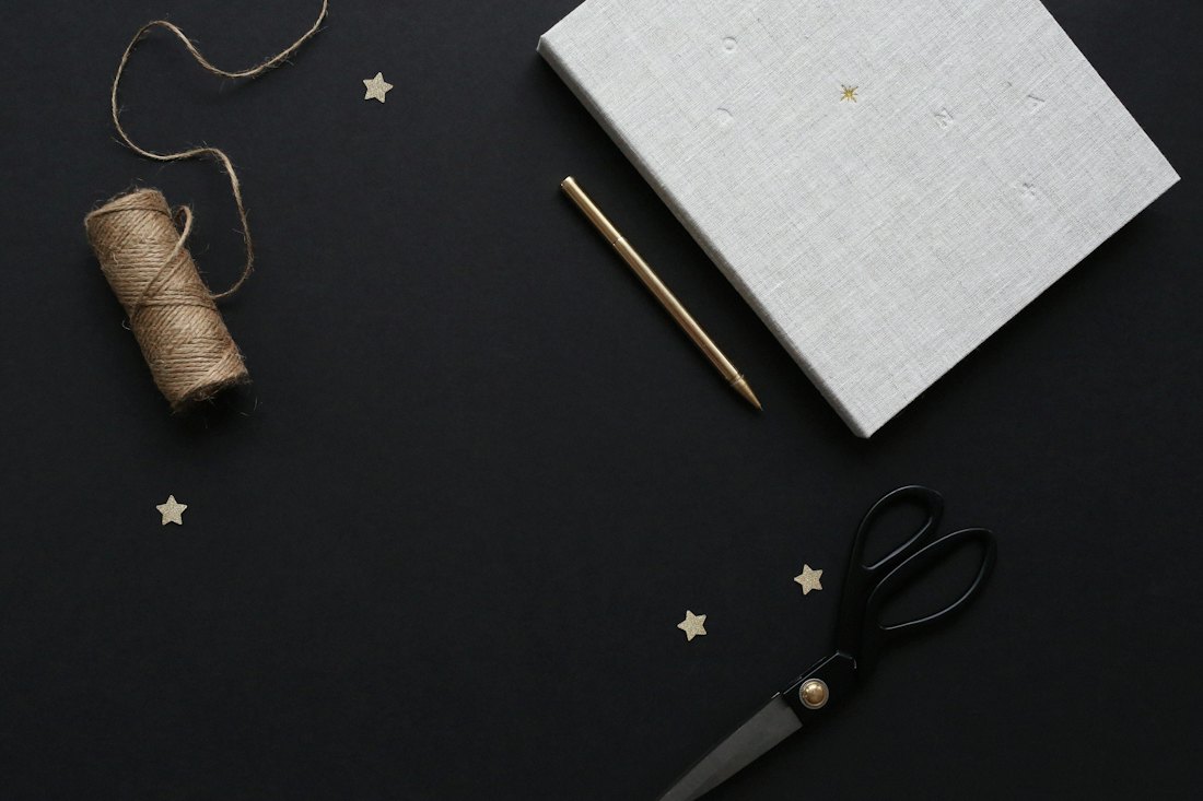 A linen-covered notebook, a pencil, a spool of twine and scissors with small gold stars on a dark surface