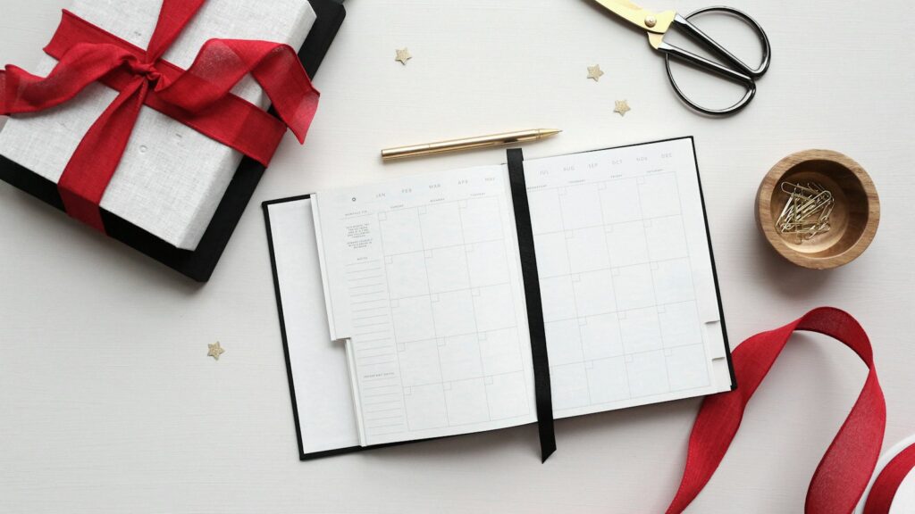 An open monthly planner beside a gold pen, scissors, red ribbon and a wrapped gift on a white desk