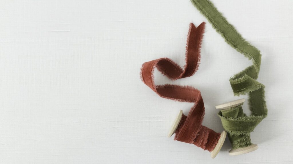 Two wooden spools of frayed silk ribbon in rust red and olive green on a pale linen background
