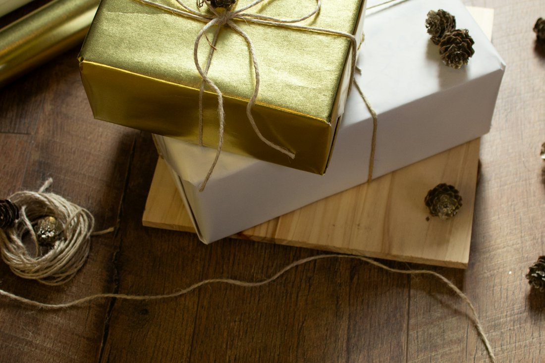 Gifts wrapped in gold and white paper tied with twine and small cones, beside rolls of wrapping paper on a wooden floor